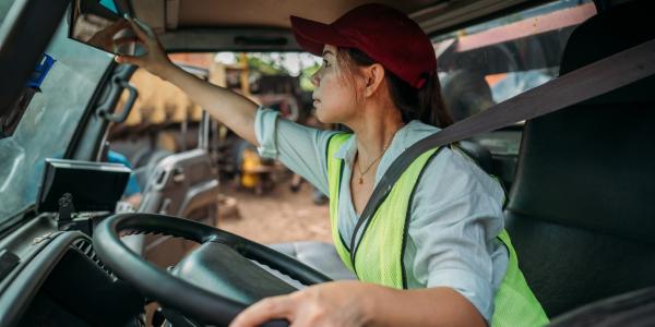 Person in commercial vehicle wearing hat and reflective vest sitting in driver’s seat, adjust mirror and looking straight ahead.