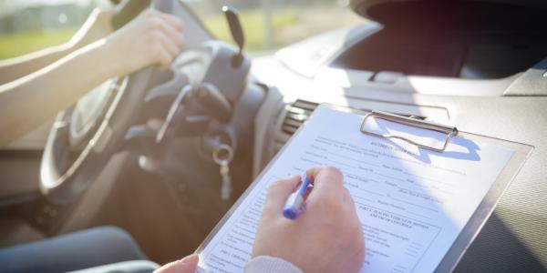 Examiner filling in a driver's license road test form sitting with their student inside a car.